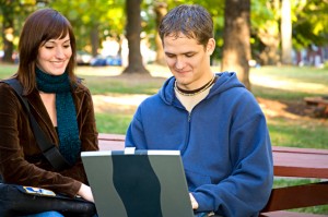 Two young adults sit on a park bench, one using a laptop.